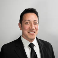 A person in a suit smiling softly at the camera, positioned against a plain gray background.