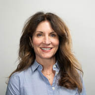 A person smiling, wearing a blue shirt, against a plain light gray background.