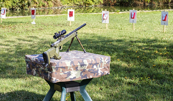 Rifle rests on camo table outdoors, aimed at numbered red targets on grassy field.