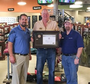 Three men stand in a store; the center man holds a framed certificate.