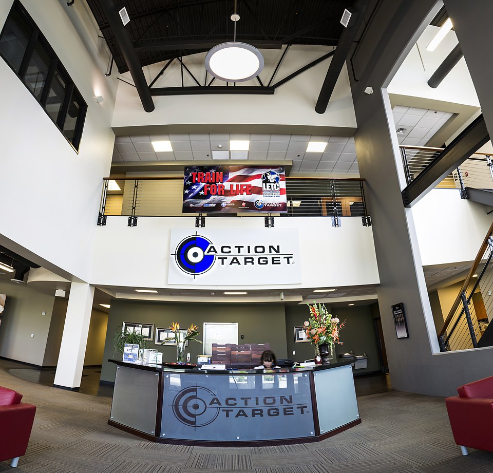 Reception desk displays "Action Target" logo; person sits behind it; office interior context. Sign above reads "TRAIN FOR LIFE LETC."