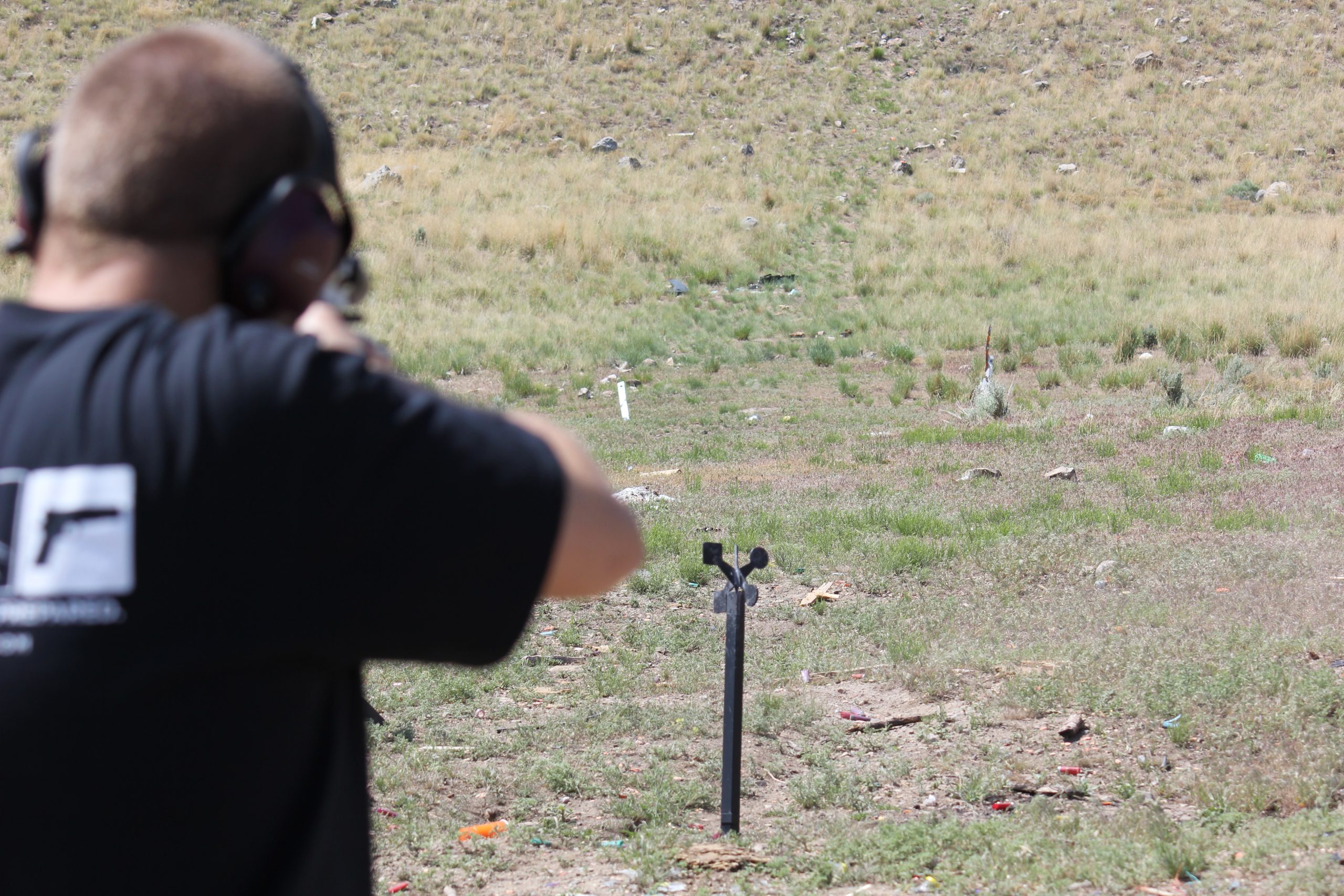 Person wearing hearing protection aiming at a steel shooting target spinner set up outdoors in a grassy open field.