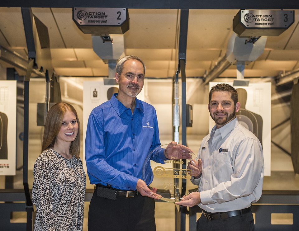 Three people hold an award in a shooting range; targets visible in background.