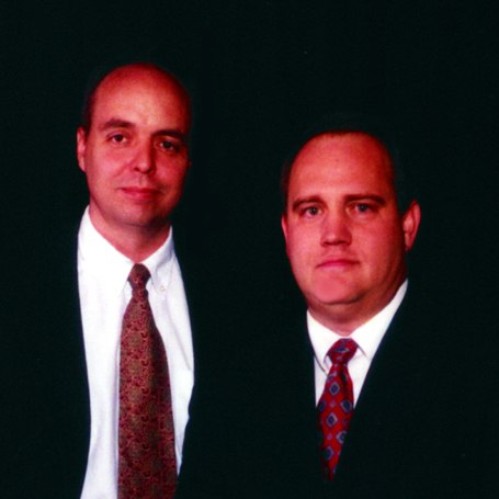 Two men in business suits and patterned ties posing together against a dark background for a formal portrait.