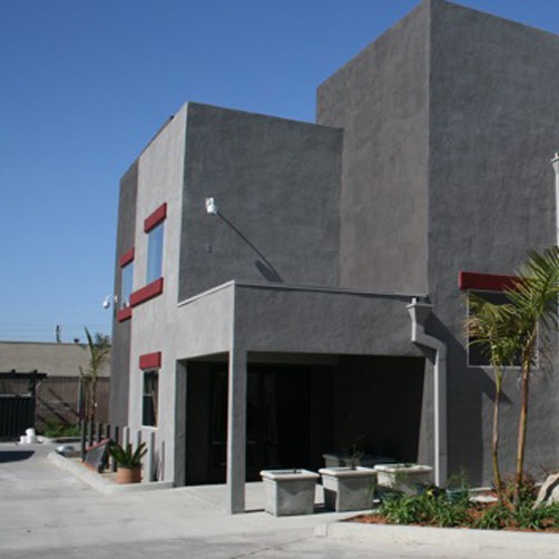 Modern gray two-story building with red window accents, flat roof, and landscaped entrance with small palm trees.