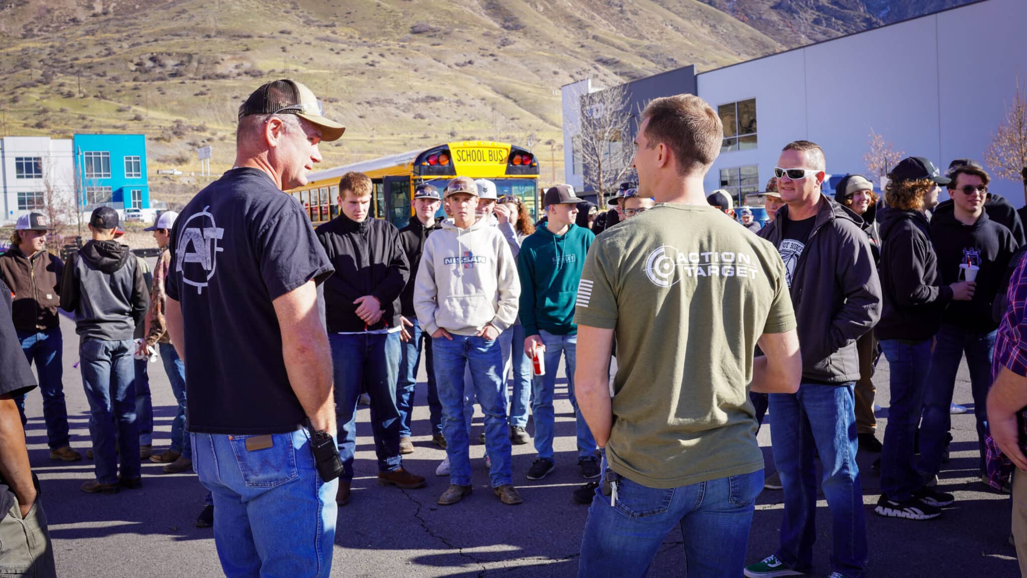 Group of people in casual clothes and Action Target shirts gathered outside near a school bus.