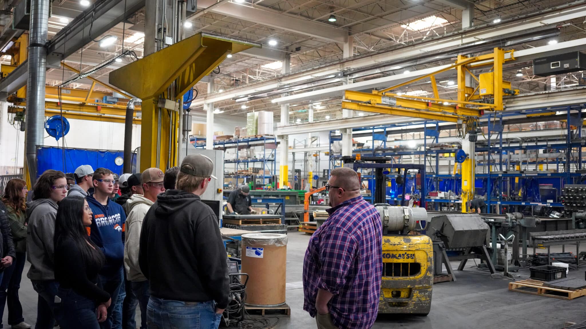 People standing in a factory tour, observing machinery and workers in a large industrial space.