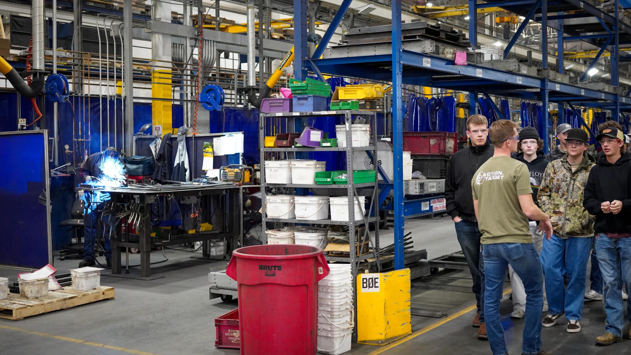 Welder works at a bench in a busy factory, while a group tours nearby.