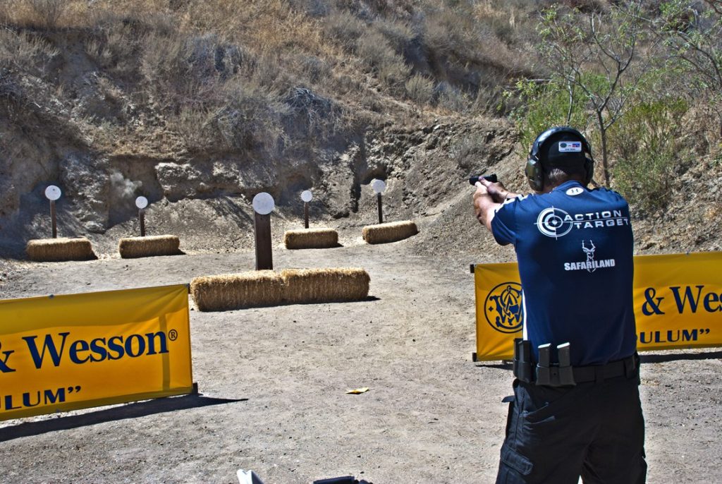 Man wearing hearing protection and Action Target shirt aiming a handgun at steel plate targets during an outdoor shooting competition.