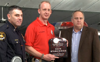 Three men, including a police officer, stand together as one presents an award plaque.