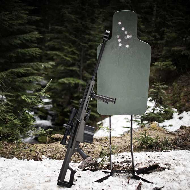 Green steel silhouette target outdoors with rifle leaning against it, showing bullet impacts on upper area.