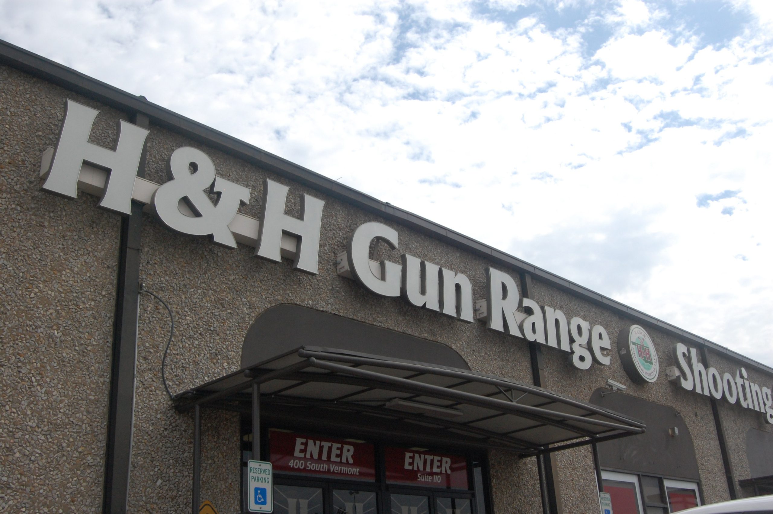 Sign for "H&H Gun Range Shooting" on a stone building facade under a cloudy sky.