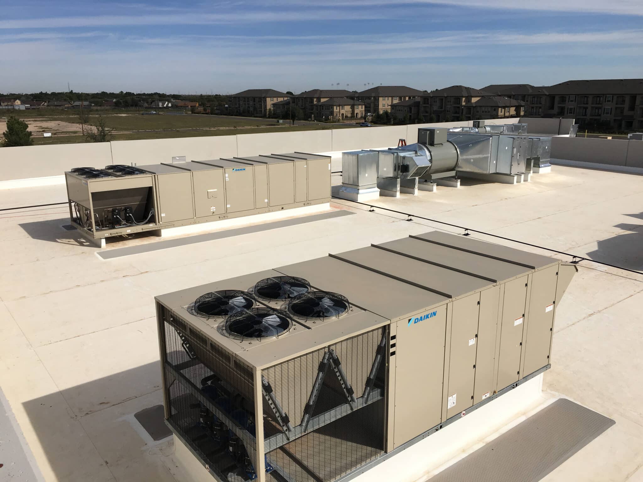 Large HVAC units on top of a building roof with a field and housing in the backdrop.