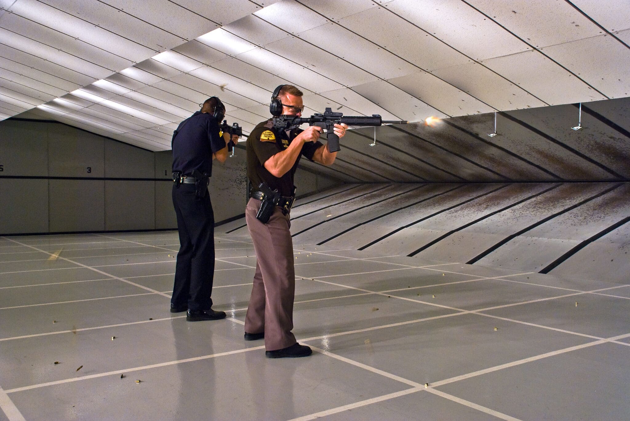 Two officers are firing rifles at targets in an indoor shooting range.