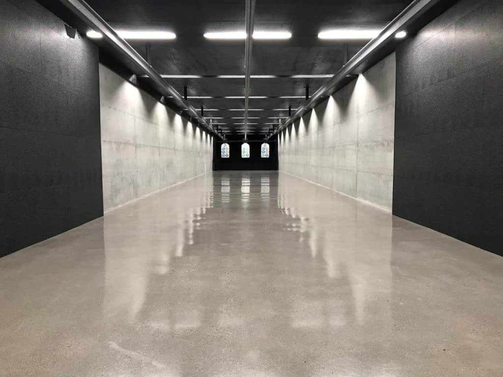 Indoor shooting range with polished concrete floors, gray walls, overhead lighting, and distant paper targets at the end of the lanes.