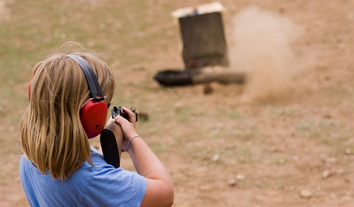 Person wearing ear protection aims a rifle at a dusty target outdoors.