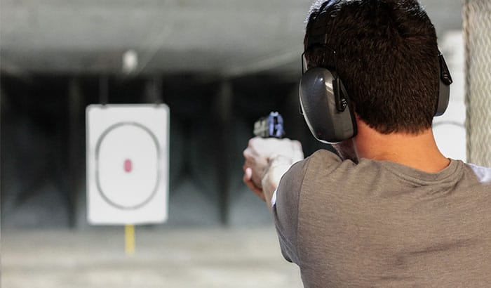 Man wearing hearing protection aiming a handgun at a paper target inside an indoor shooting range.