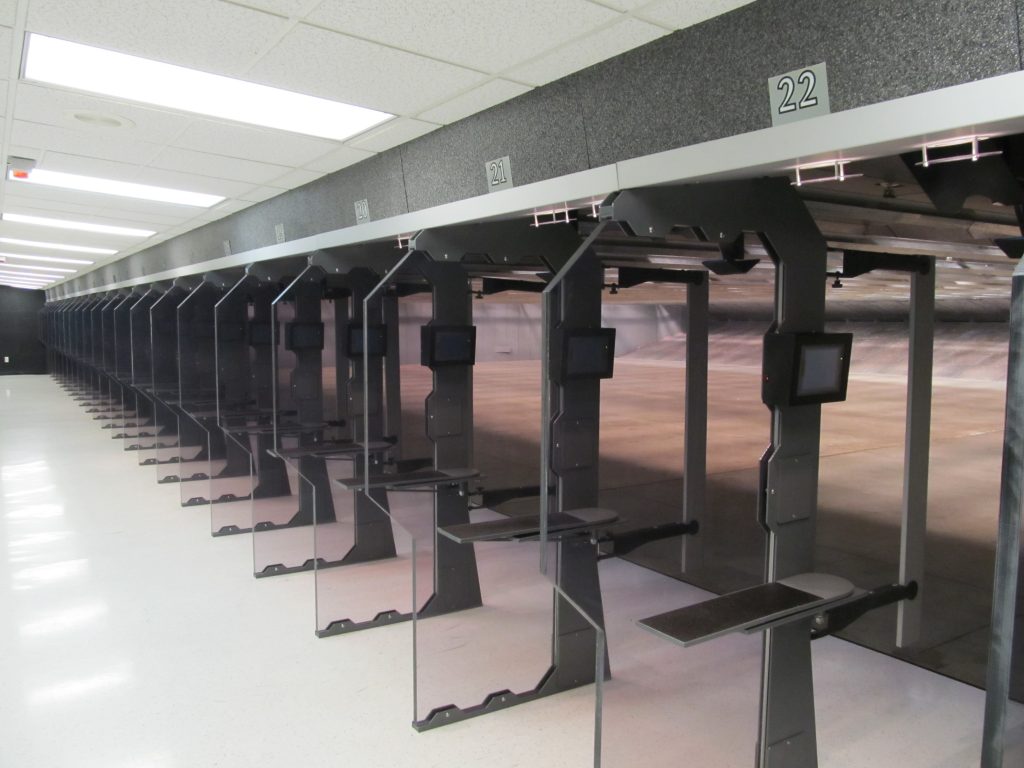 Row of indoor shooting range stalls with numbered lanes, glass dividers, and digital control screens at each station.