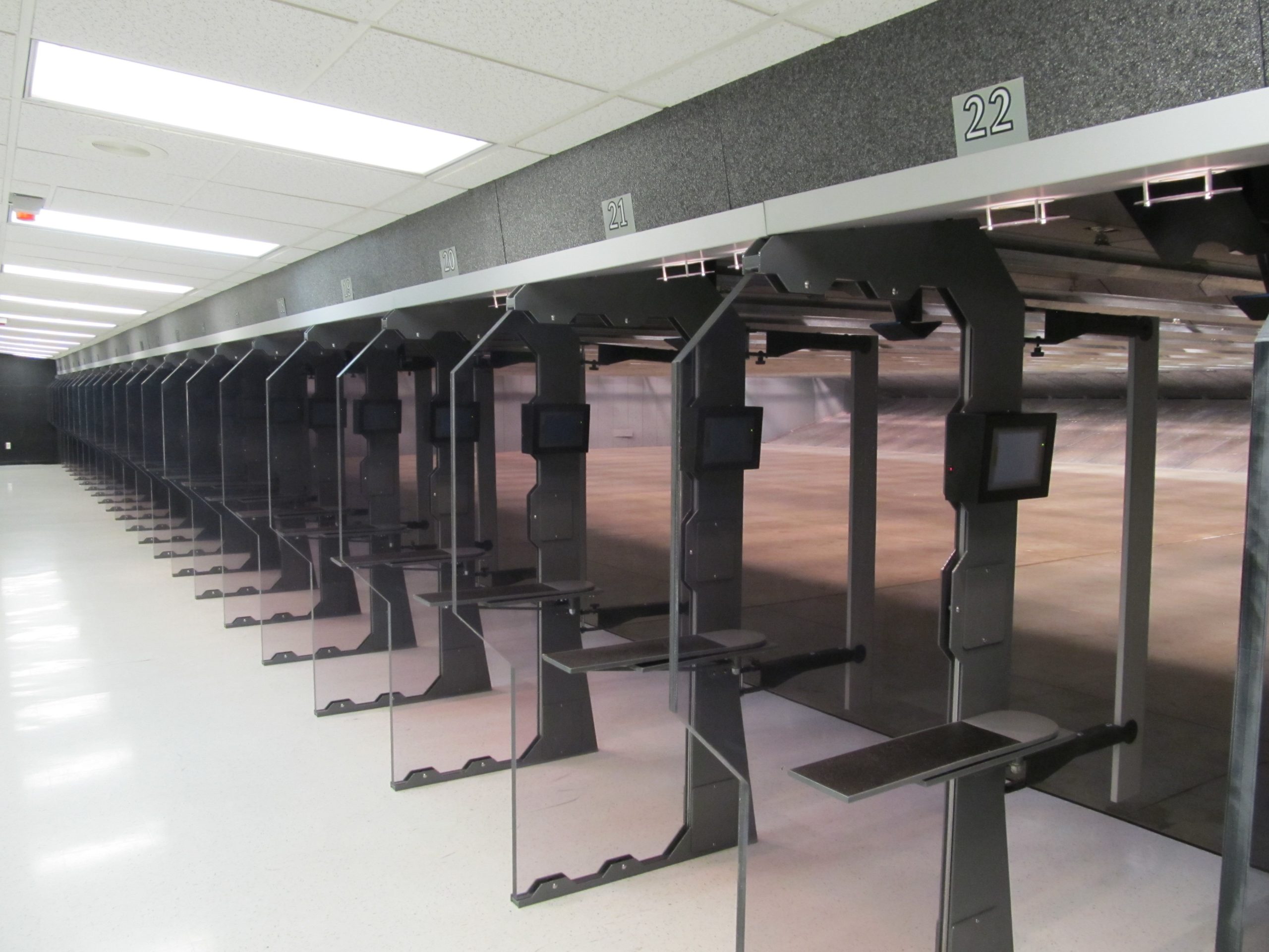 Row of indoor shooting range stalls with numbered lanes, glass dividers, and digital control screens at each station.