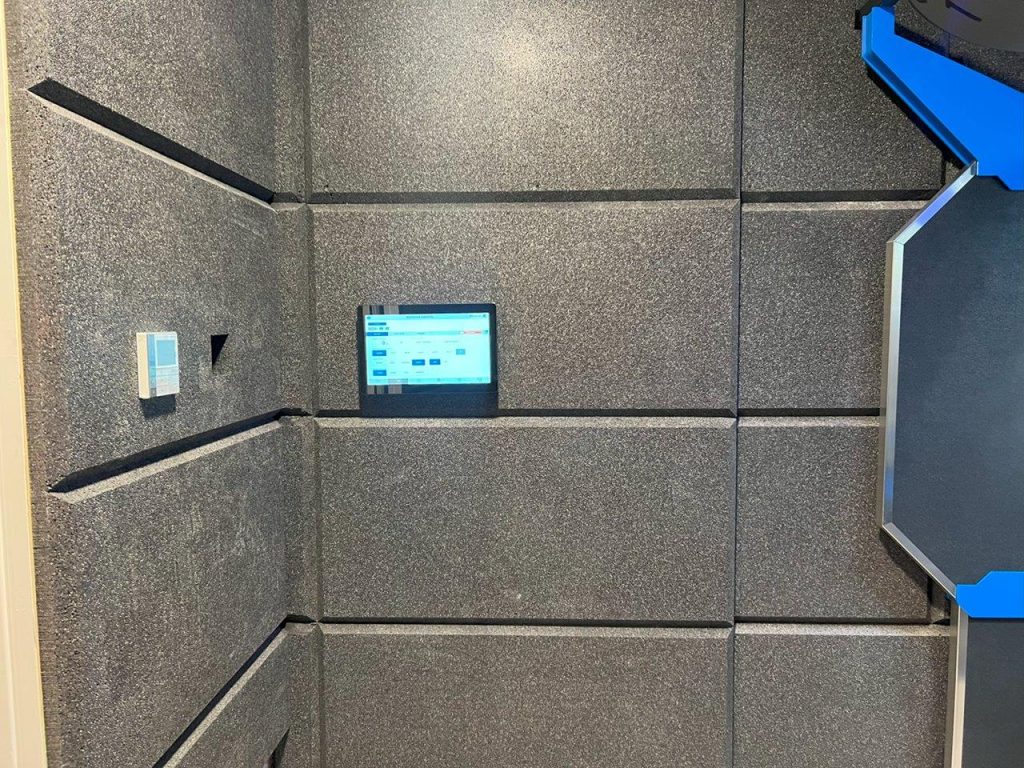 Tablet displaying controls on gray acoustically-treated wall; adjacent blue panel.