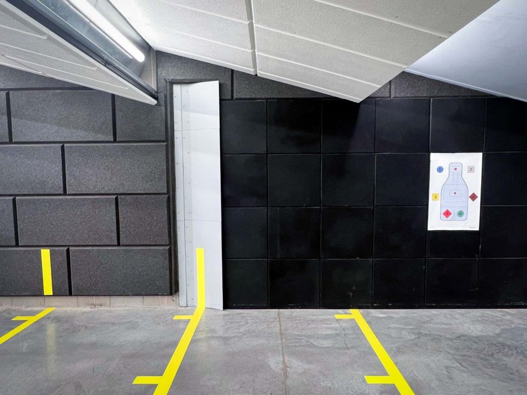 Indoor shooting range wall with black ballistic panels, yellow safety lines, and paper target mounted for training.