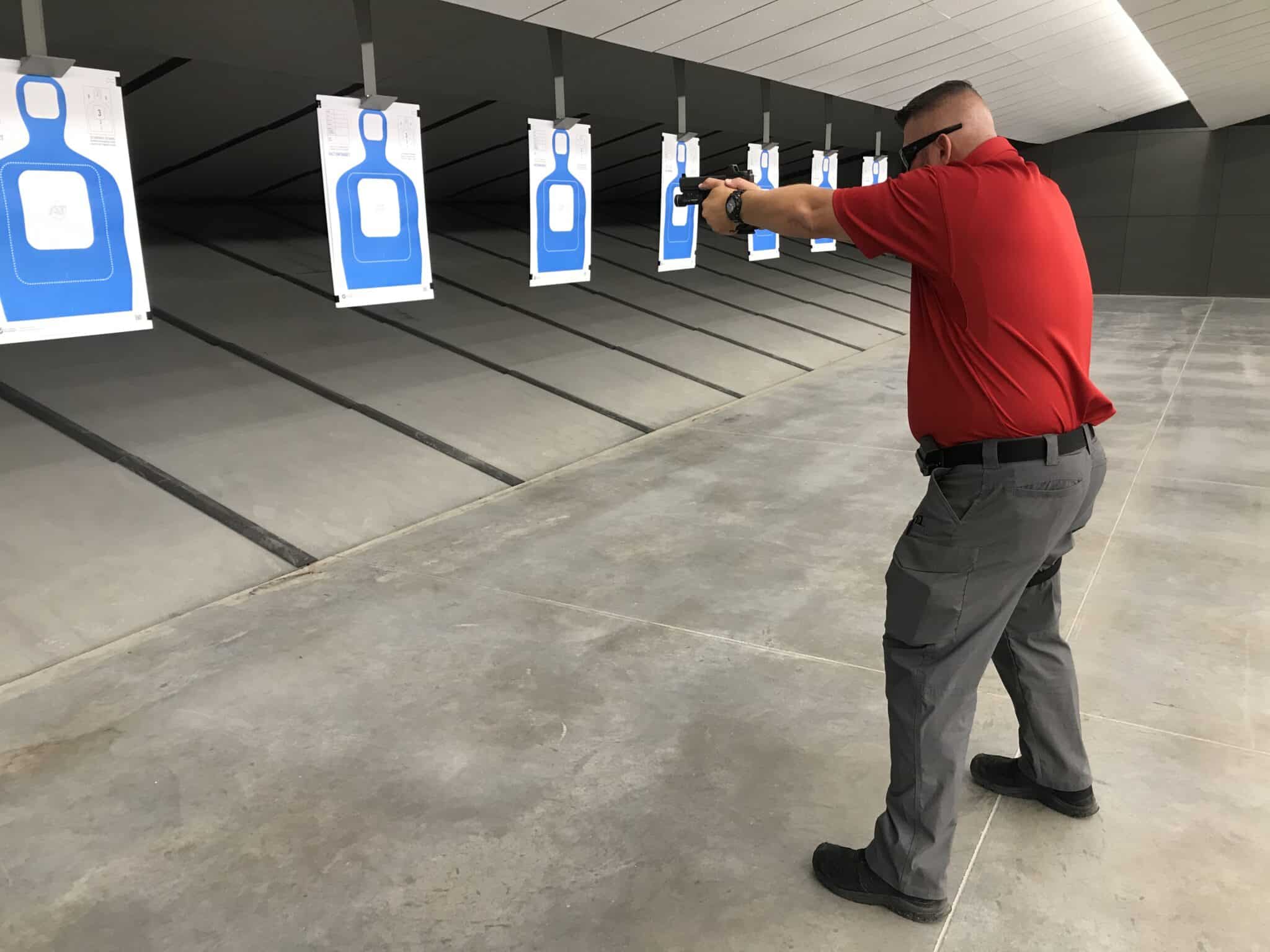 Man in red shirt aiming a handgun inside an indoor shooting range with blue silhouette paper targets on the lanes.