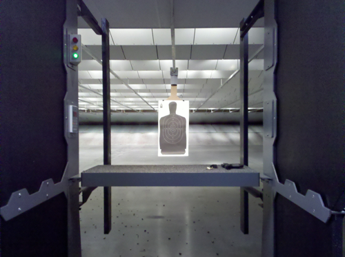 Indoor shooting range lane with control panel, bench, and silhouette paper target suspended downrange.