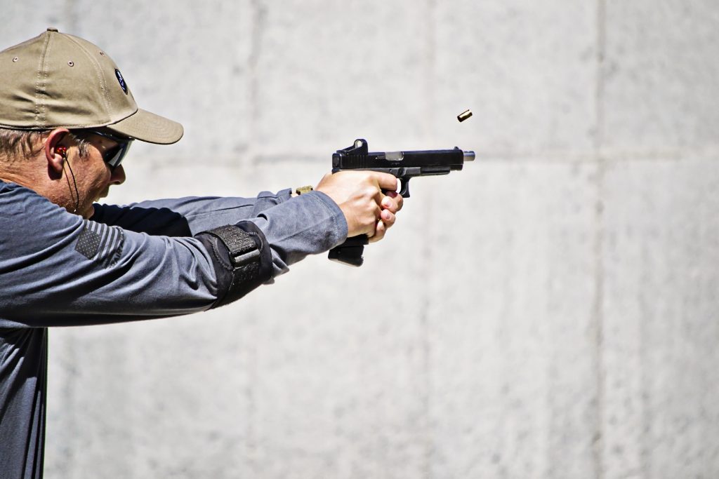 Person firing handgun, ejecting casing; wearing cap, glasses, in an outdoor shooting range.