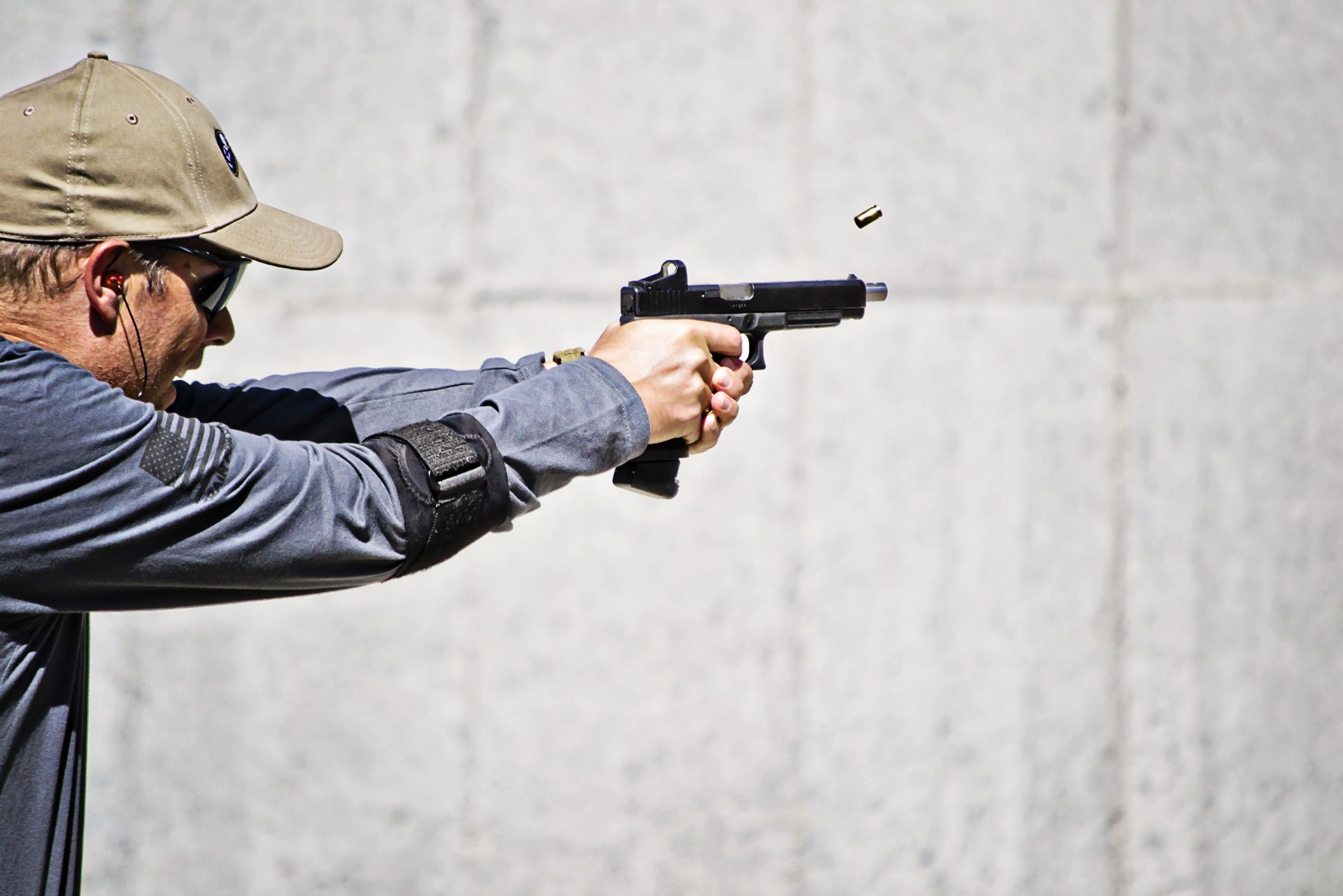 Person firing handgun, ejecting casing; wearing cap, glasses, in an outdoor shooting range.