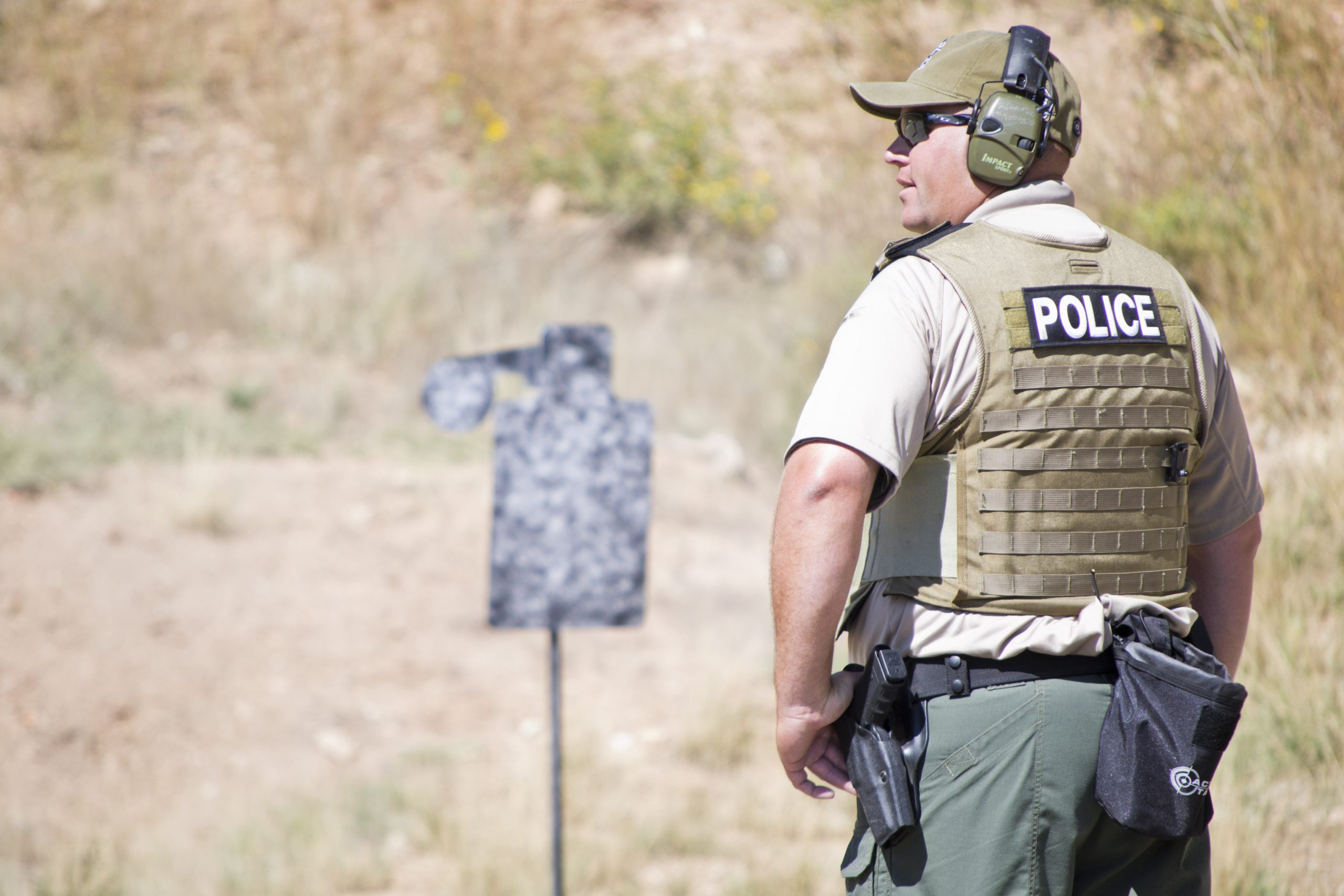 Police officer standing near firearm target; wearing tactical gear outdoors.