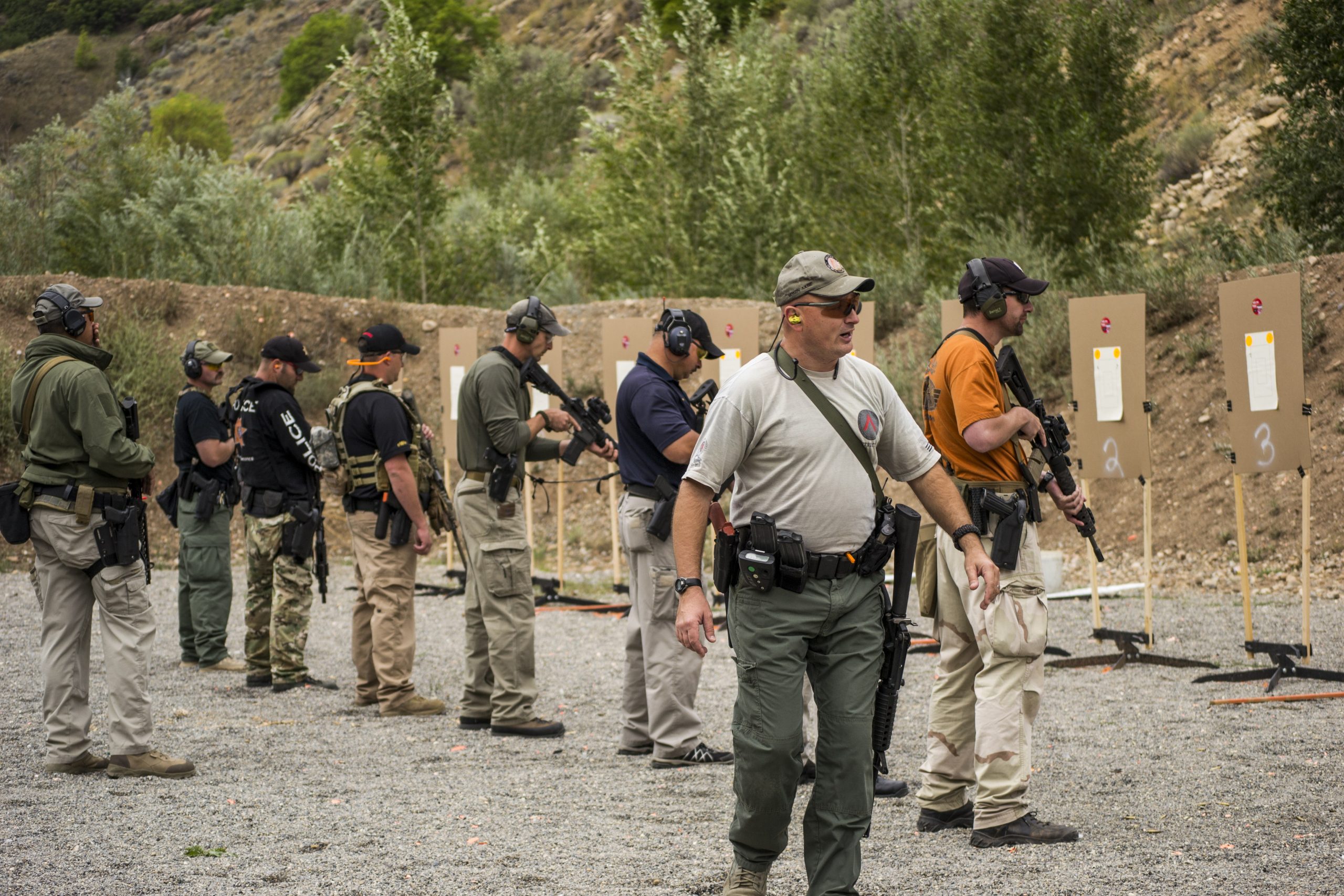 Instructors and trainees hold guns, stand by targets on a gravel shooting range with trees around.