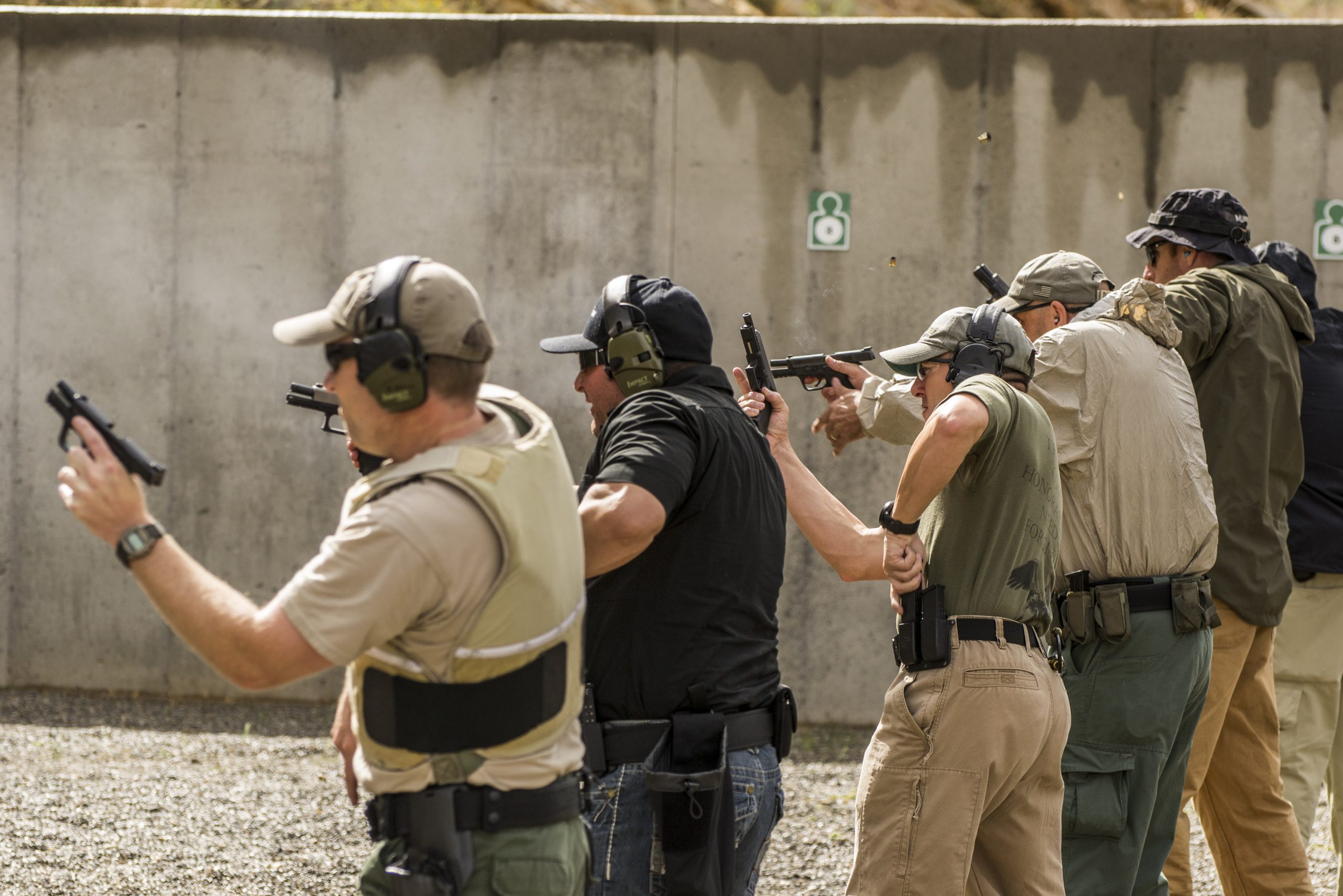 Group shooting handguns, wearing ear protection, at an outdoor firing range, concrete wall background.