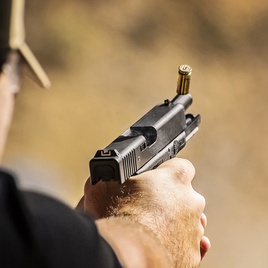 Hand holding a pistol, ejecting a bullet casing, outdoors with blurred background.