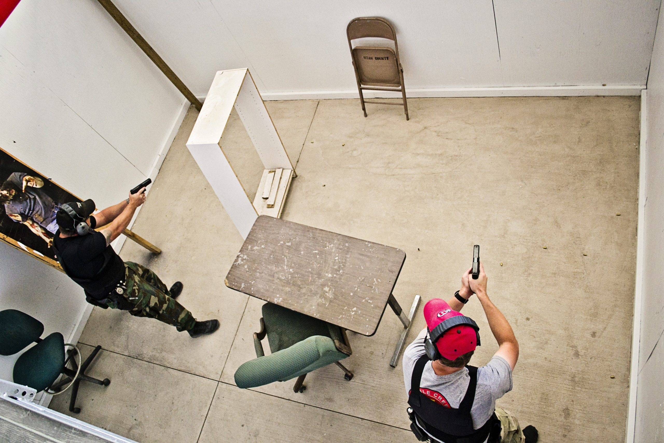 Two people in a shoot house training scenario aiming pistols, with chairs and a table set up as obstacles.