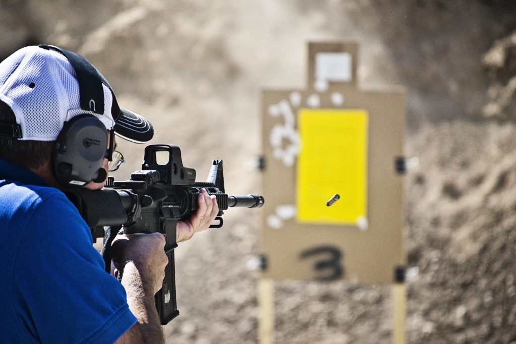 Man firing a rifle at a cardboard target on an outdoor shooting range.