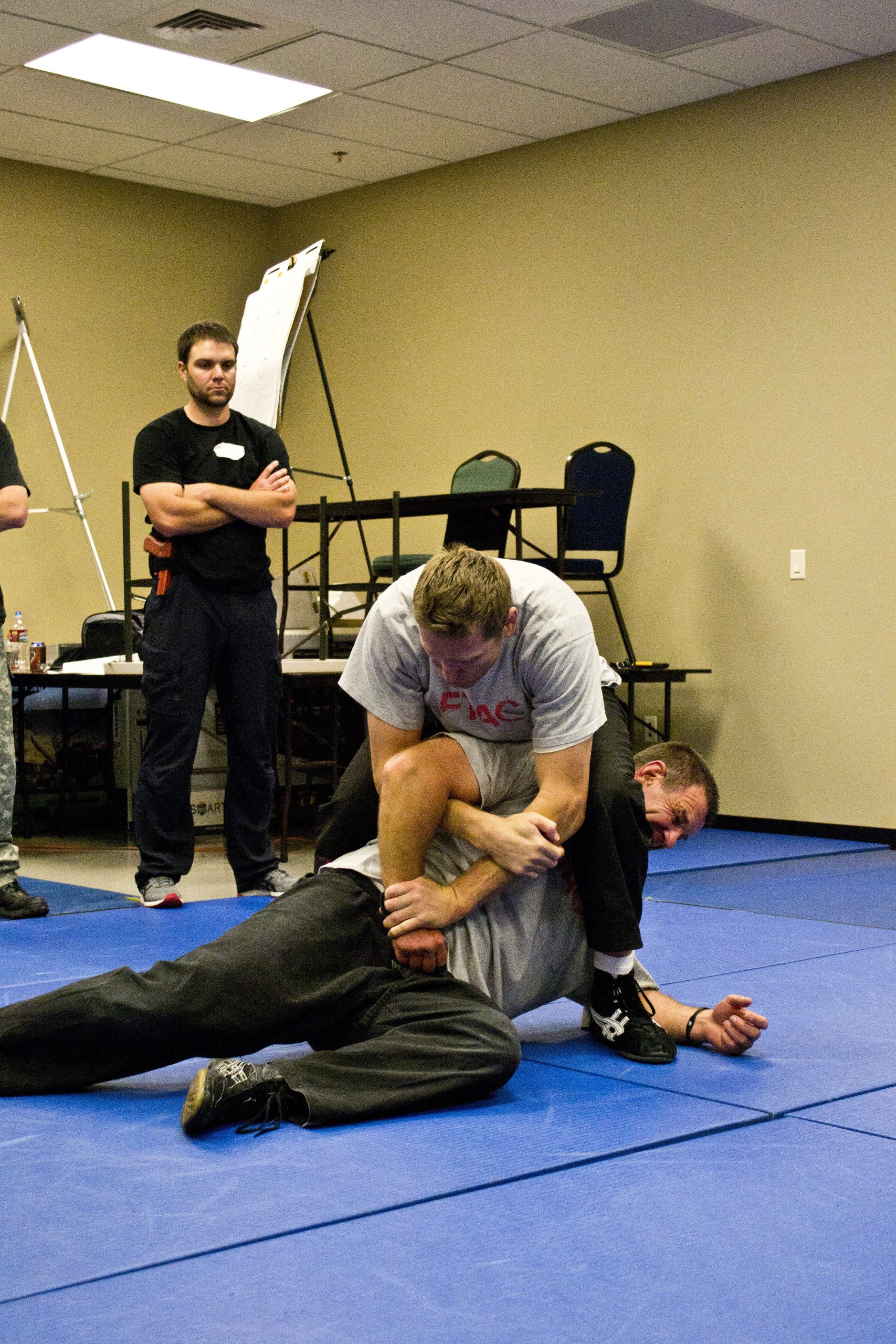 Person restraining another on a blue mat; others watch in a training room.