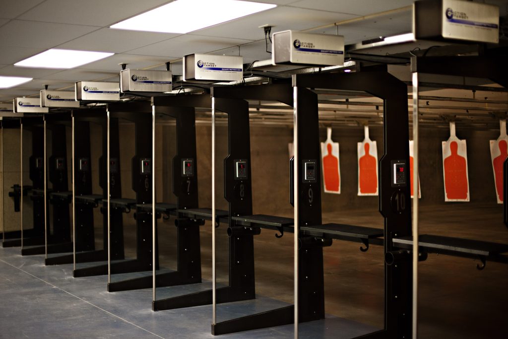 Row of indoor shooting range stalls with electronic controls and orange silhouette paper targets downrange.