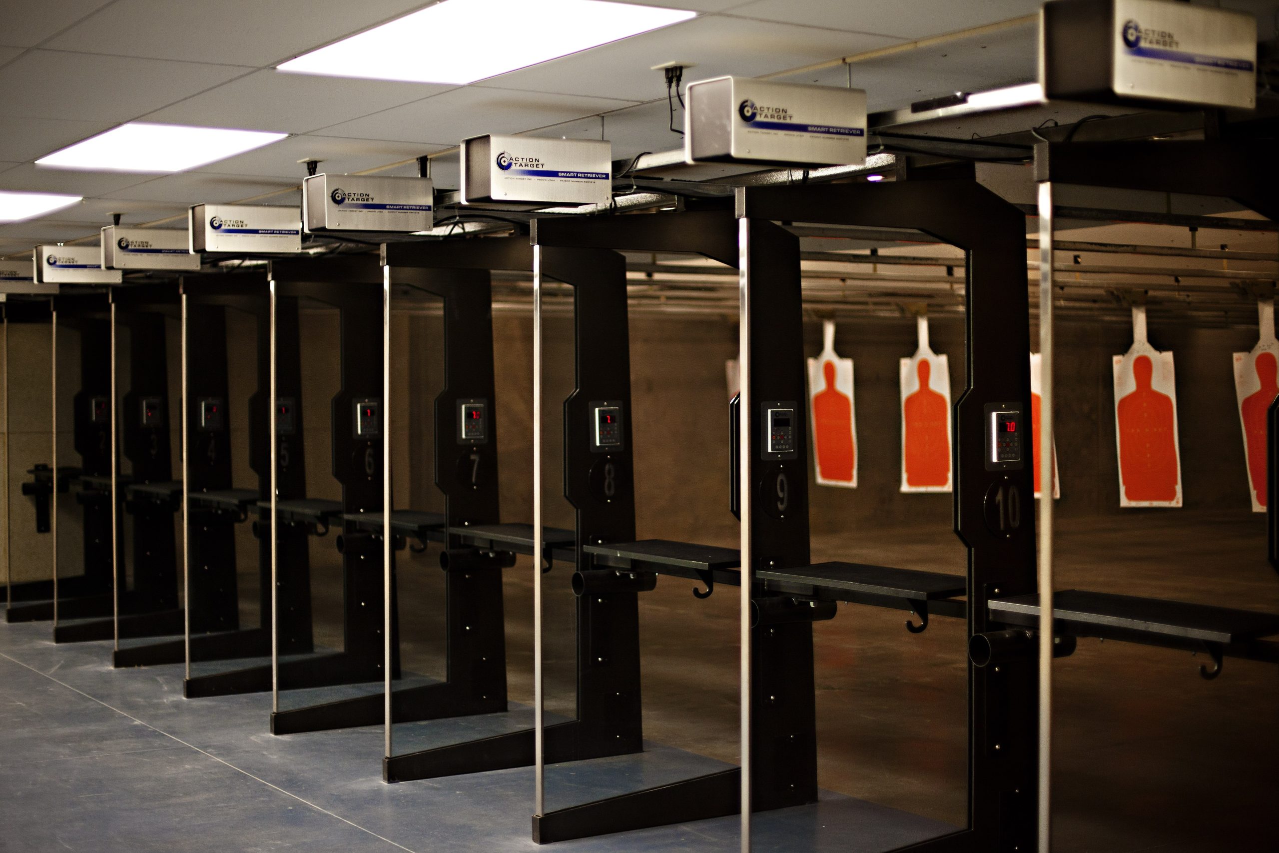 Row of indoor shooting range stalls with electronic controls and orange silhouette paper targets downrange.