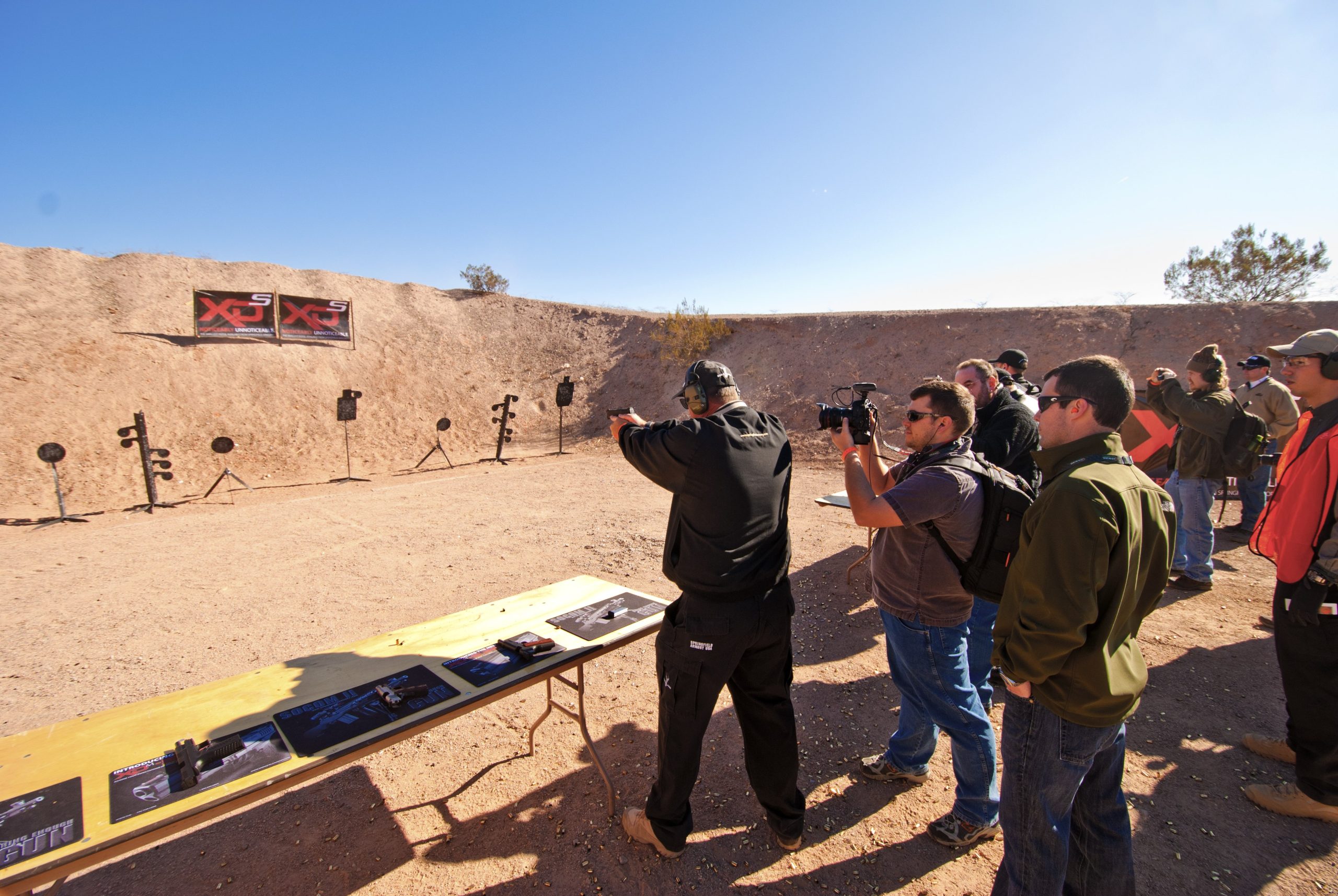 Group of people at an outdoor shooting range observing a man firing a handgun at steel targets.