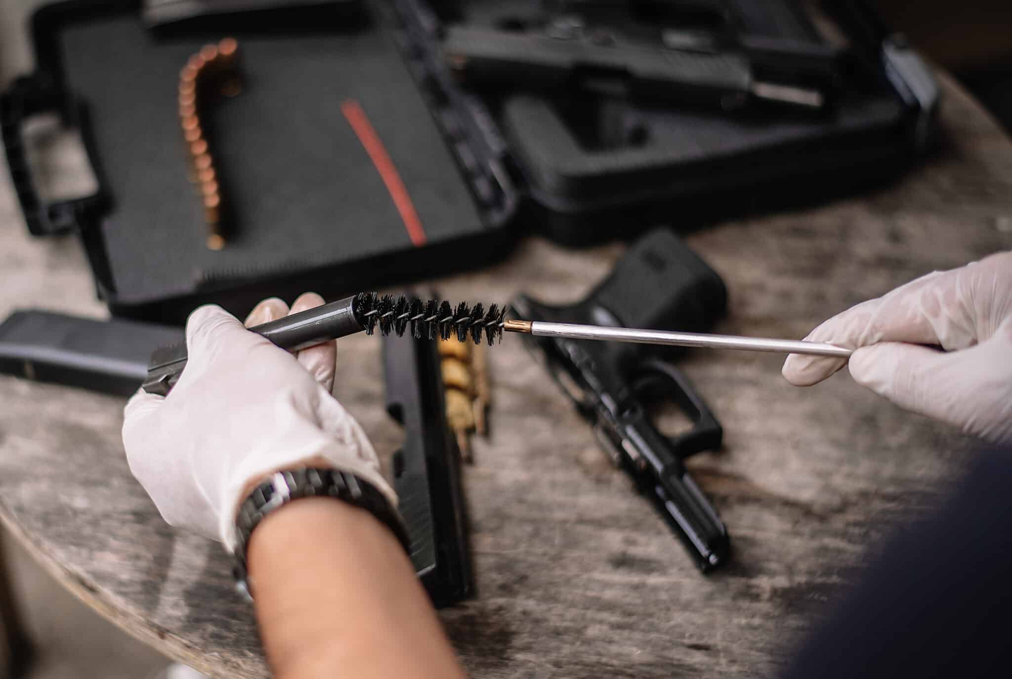 Person wearing gloves cleaning a handgun barrel with a bore brush, with firearm parts and ammo visible on the table.