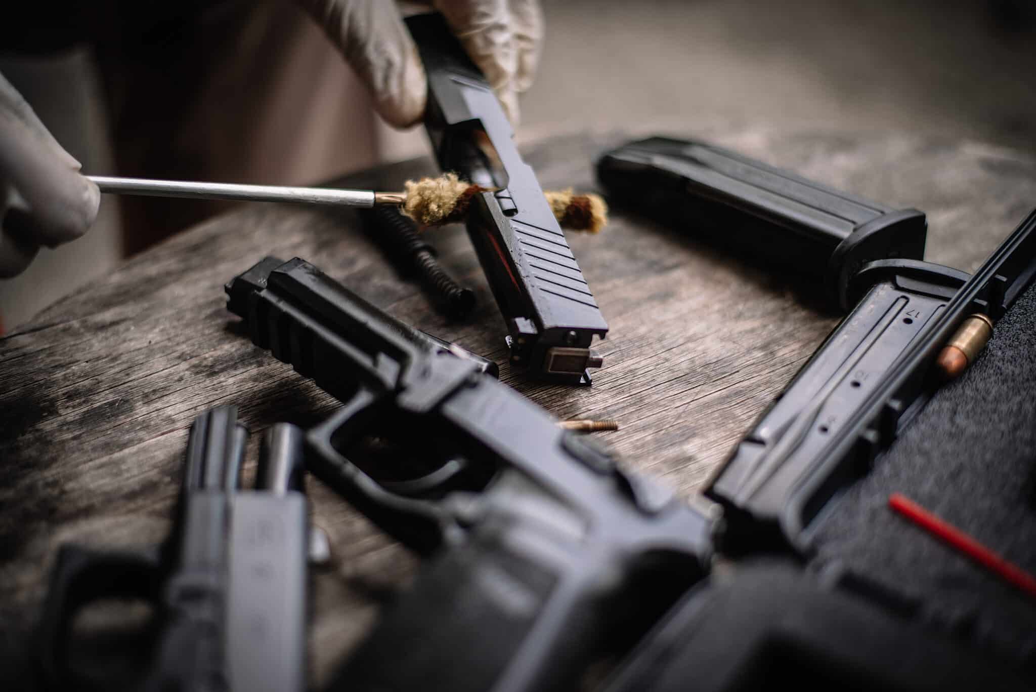 Close-up of a person cleaning a disassembled handgun with a cleaning rod, showing pistol parts, magazines, and ammunition on a wooden surface.