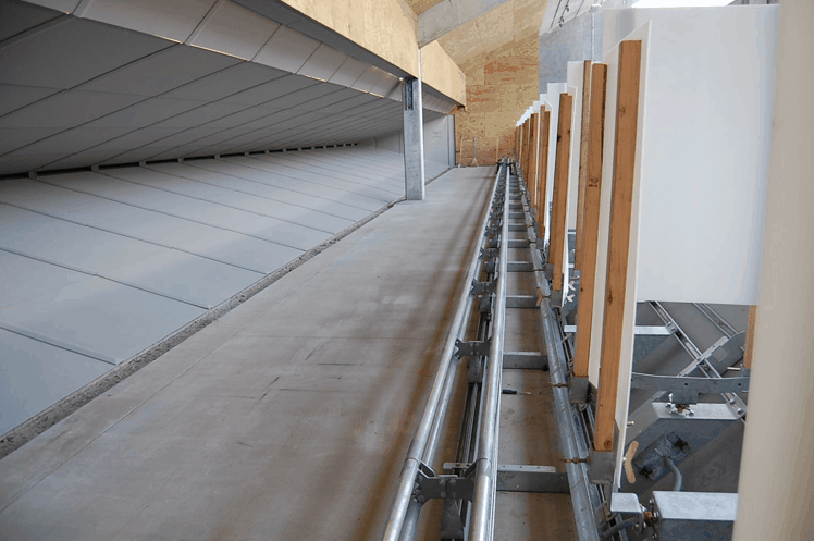 Metal pipes run parallel to a concrete walkway in an industrial building with a slanted roof.