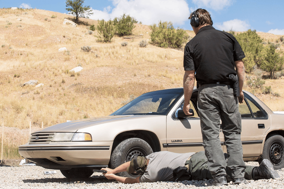 A man lies by a car labeled "Lumina," aiming a weapon. Another stands nearby in a grassy area.