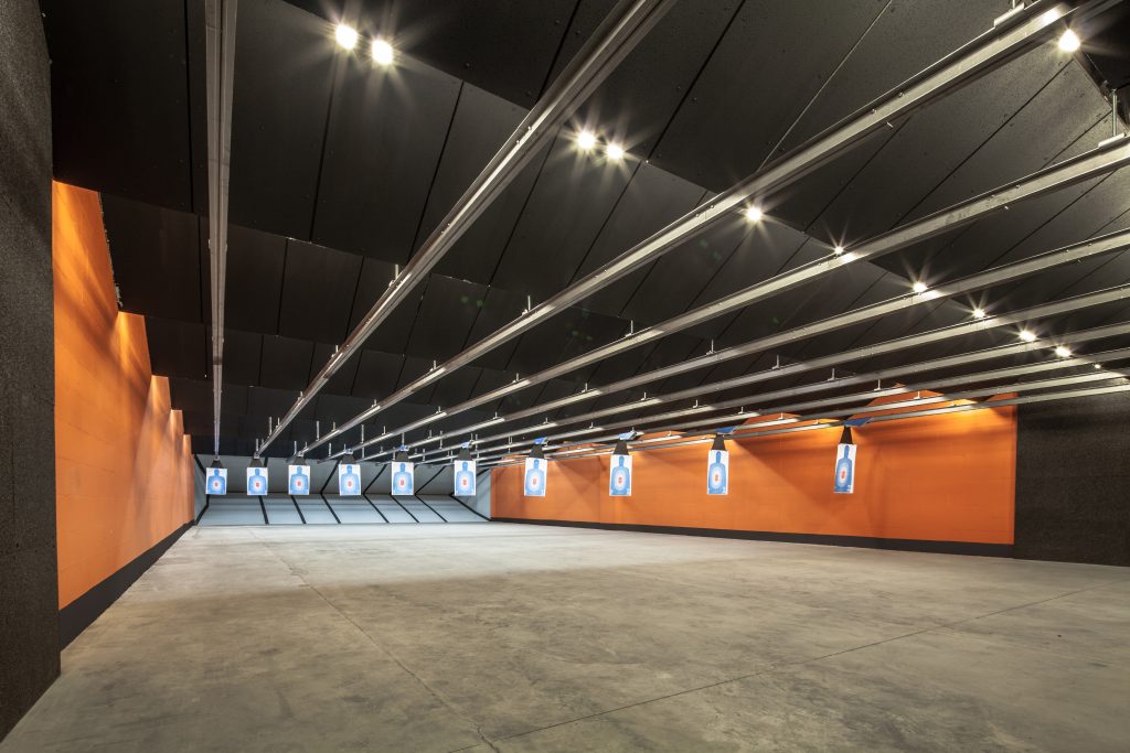 Shooting range with target sheets hanging from railings, bright lighting, and orange walls.