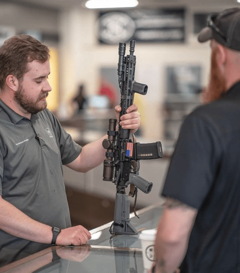 Man holding a rifle vertically, standing behind a glass display counter, talking to another man.