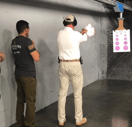 A person fires a gun at a target inside a shooting range, with another person observing.