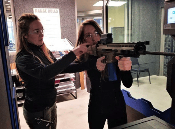 Two people at a shooting range; one instructs the other on holding a rifle. 