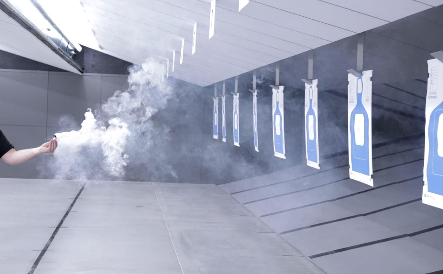 Person releasing smoke inside an indoor shooting range with blue silhouette paper targets hanging in multiple lanes.