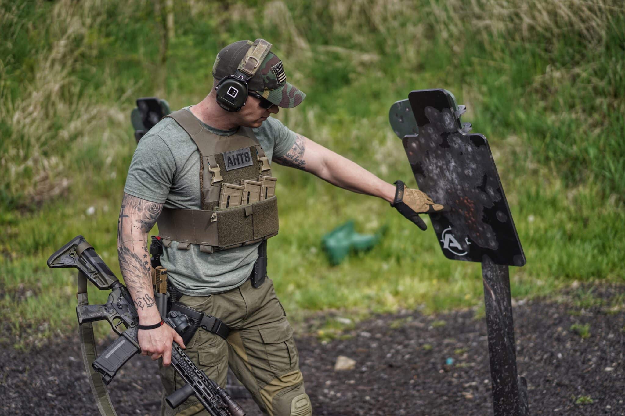 Man in tactical gear and hearing protection inspecting bullet impacts on a steel target while holding a rifle outdoors.