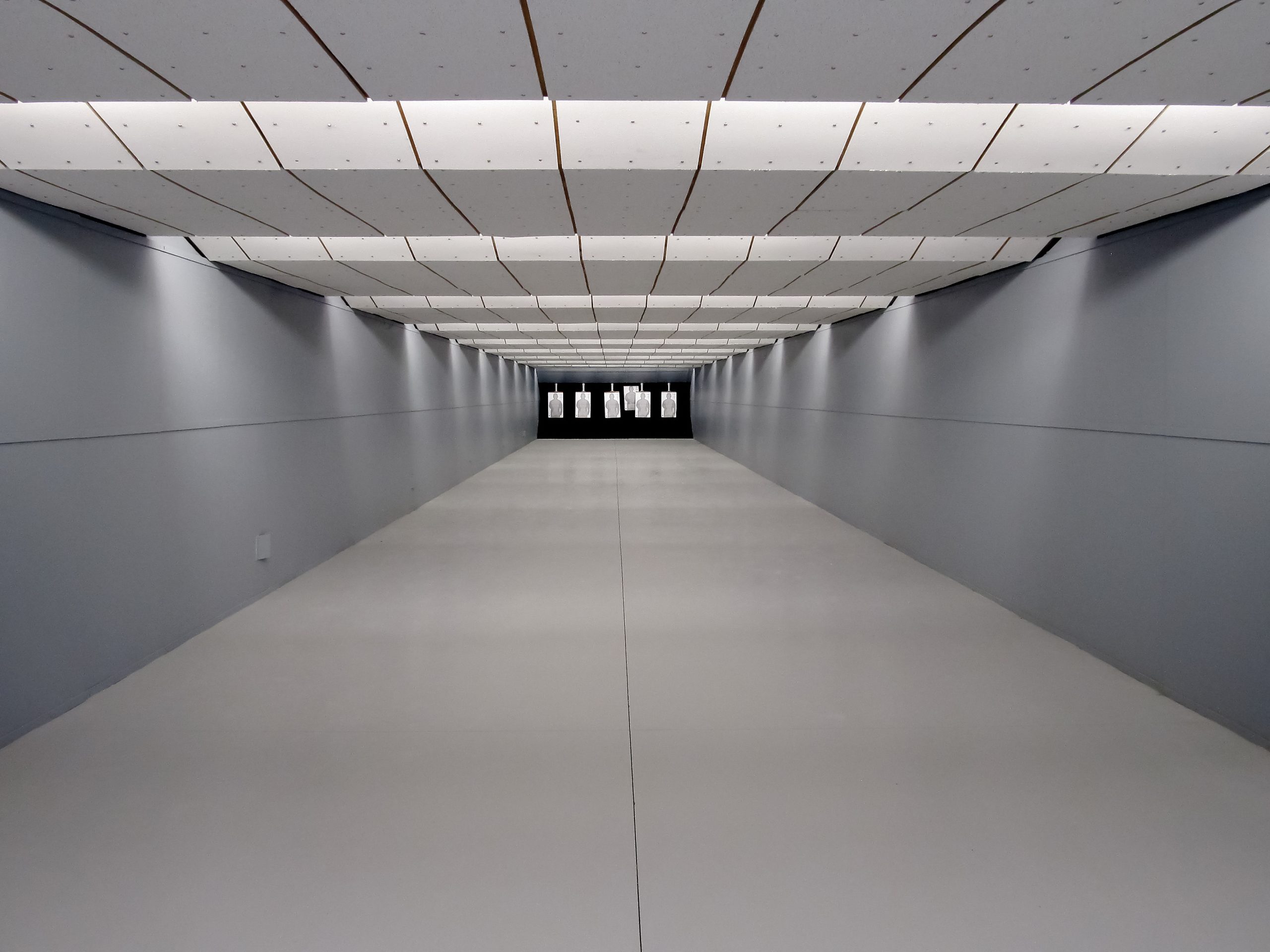 Indoor shooting range with gray walls, white acoustic ceiling panels, and paper targets positioned at the far end.